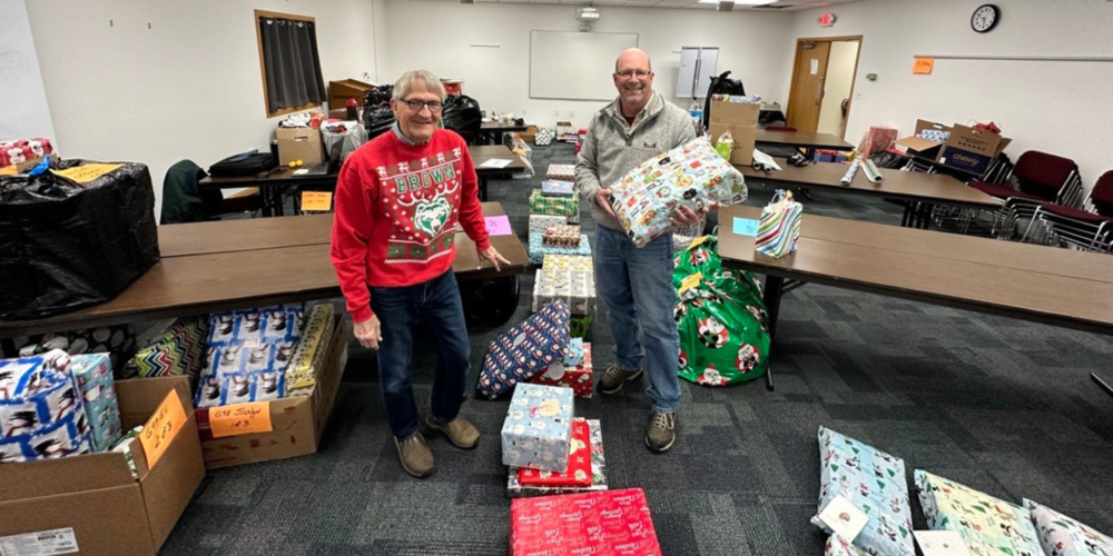Two men in holiday sweaters surrounded by boxes of brightly wrapped Christmas presents